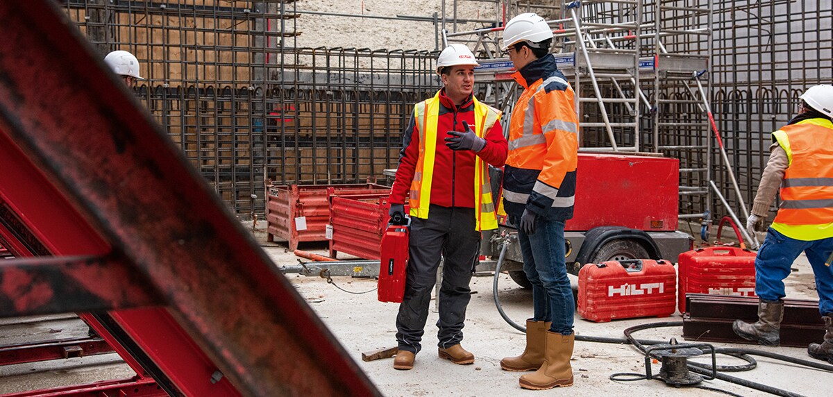 two men wearing hard hats on a construction site looking at a te 6-a cordless rotary hammer drill