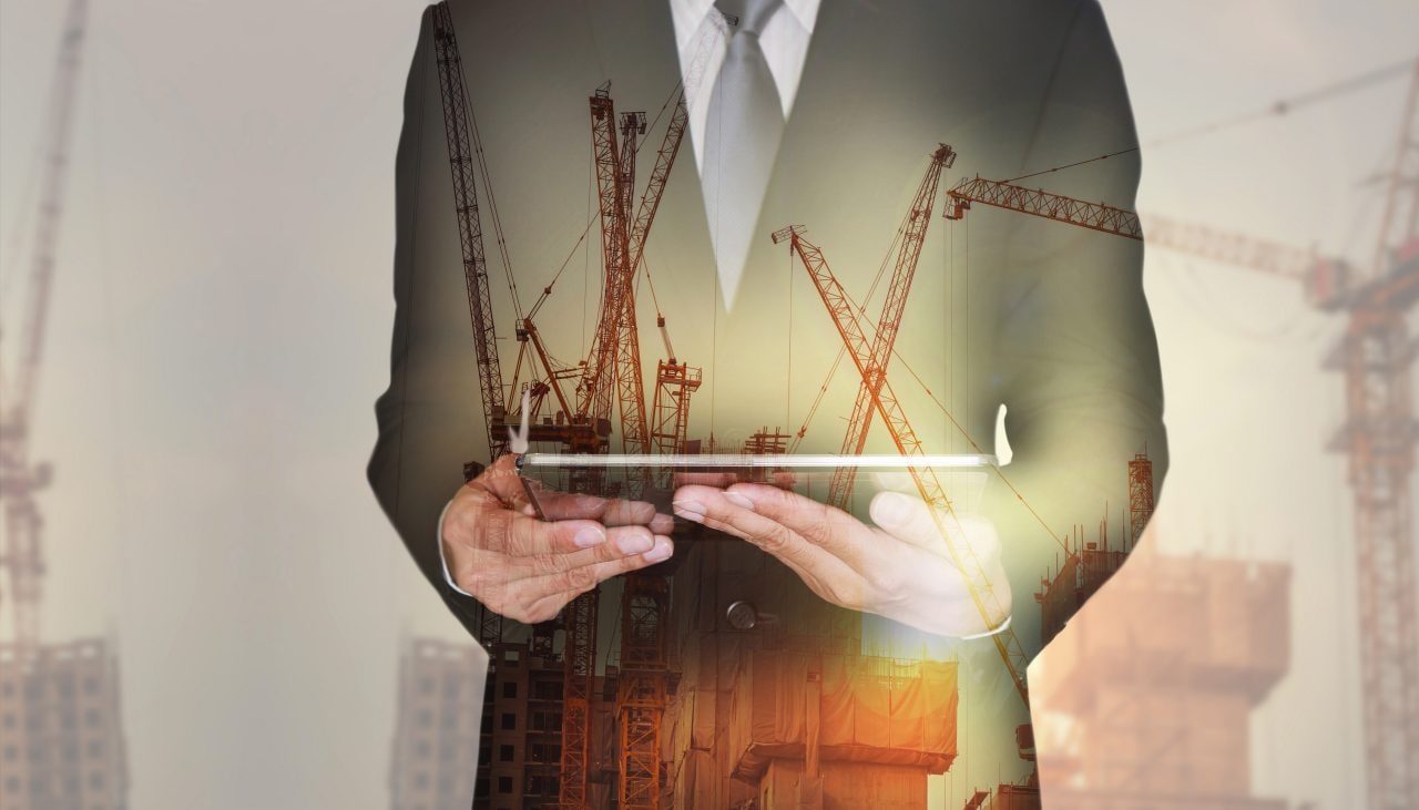 665721386 Double exposure of businessman hand hold tablet, construction crane, building and sunset in the evening as business, technology, communication and industry concept.