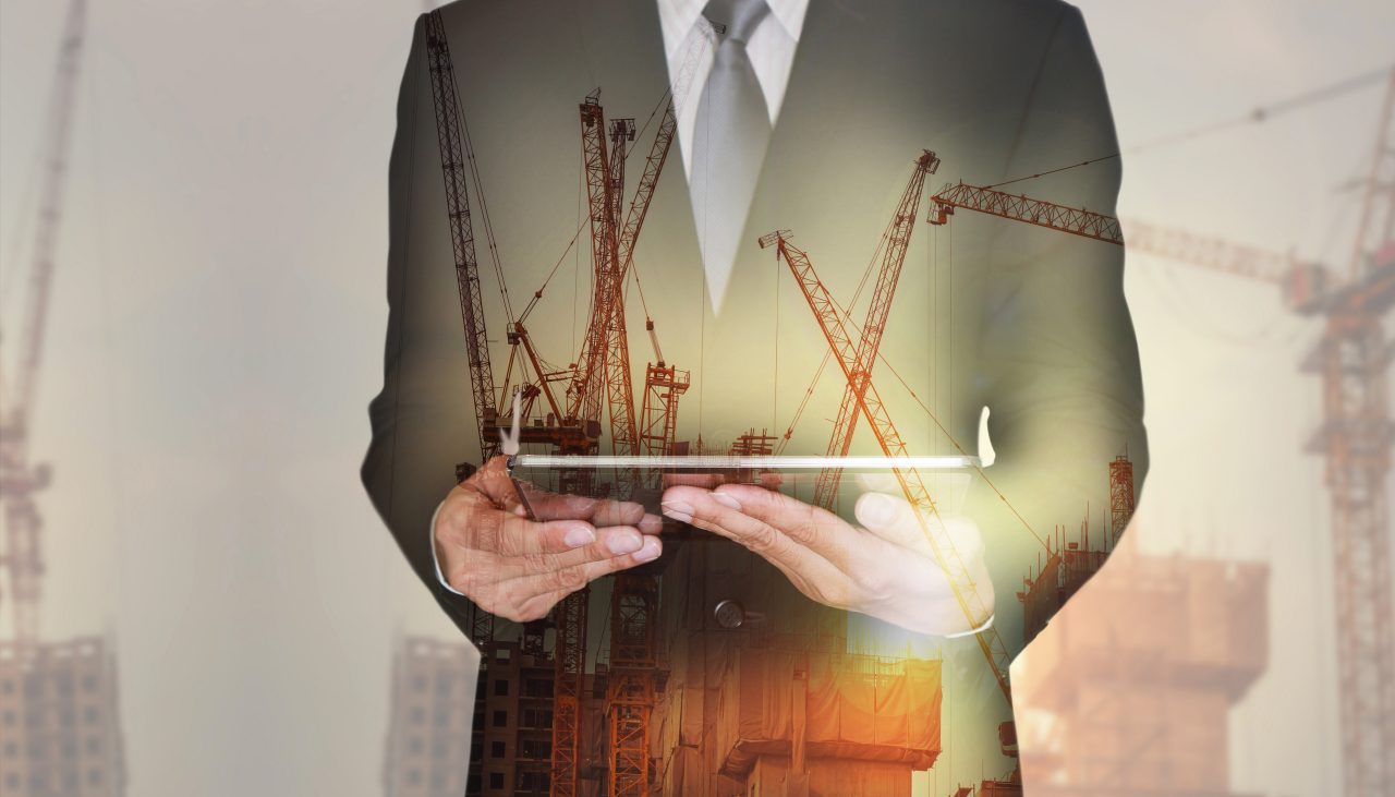 665721386 Double exposure of businessman hand hold tablet, construction crane, building and sunset in the evening as business, technology, communication and industry concept.